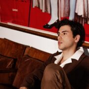 Uomo con outfit total brown seduto su una sedia vintage in un ambiente retrò. Indossa un blazer doppiopetto marrone, pantaloni coordinati e mocassini in pelle senza calze. Completa il look con occhiali da sole con lenti color ambra. Sfondo con poster vintage a tema storico e arredi in legno, creando un'atmosfera sofisticata e nostalgica.