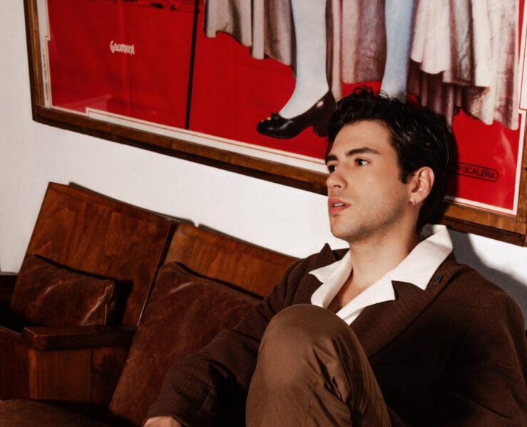 Uomo con outfit total brown seduto su una sedia vintage in un ambiente retrò. Indossa un blazer doppiopetto marrone, pantaloni coordinati e mocassini in pelle senza calze. Completa il look con occhiali da sole con lenti color ambra. Sfondo con poster vintage a tema storico e arredi in legno, creando un'atmosfera sofisticata e nostalgica.