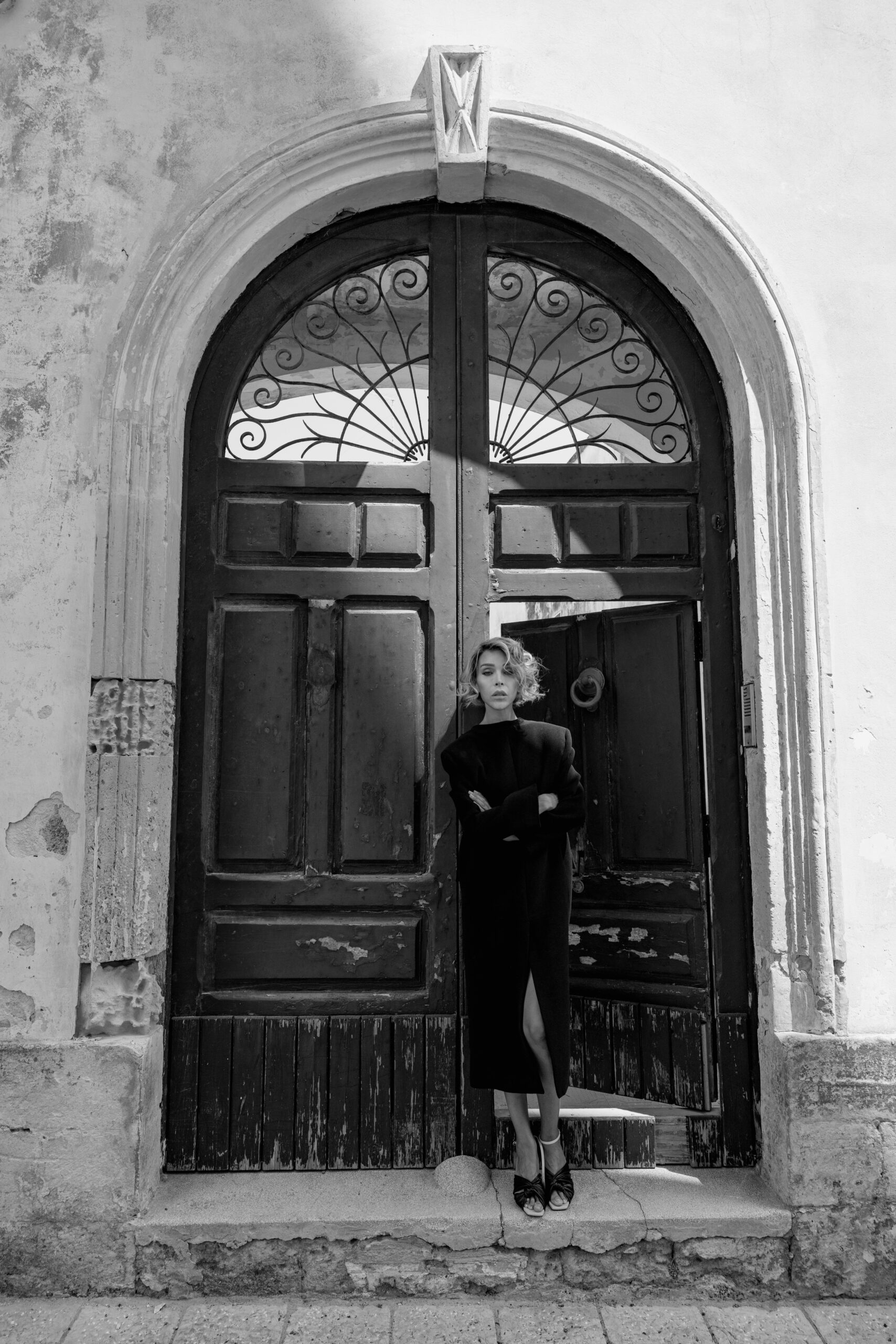 Vittoria Schisano in cappotto nero lungo posa con braccia conserte davanti a un antico portone in legno, fotografia in bianco e nero.