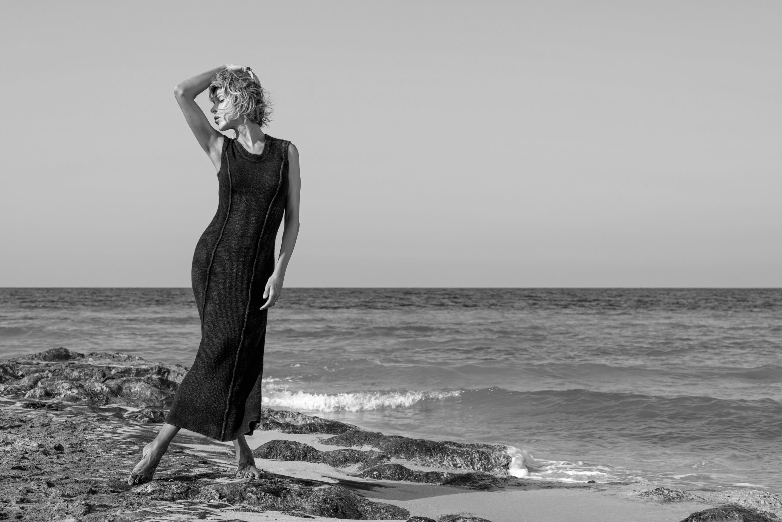 Vittoria Schisano in bianco e nero sulla riva del mare, indossa un abito Avant Toi lungo senza maniche e posa con un braccio sollevato, a piedi nudi sugli scogli